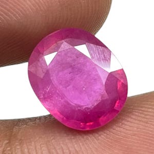 Pode incluir: Uma gema de corte oval de um tom vermelho-rosado vibrante com uma superfície facetada que reflete a luz. A pedra apresenta uma qualidade clara e transparente com uma estrutura interna visível e bordas geométricas nítidas na parte superior.