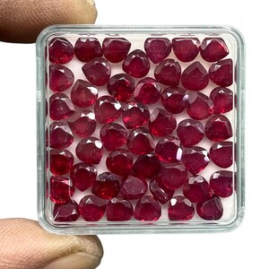 Op de afbeelding: Een doorzichtige plastic doos gevuld met talrijke gefacetteerde, dieprode edelstenen. De edelstenen zijn hartvormig en onregelmatig geslepen, met een rijke, verzadigde kleur. De doos wordt vastgehouden door de vingers van een persoon, die de edelstenen tegen een witte achtergrond toont.