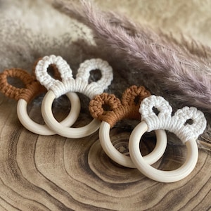 Könnte beinhalten: Drei Holz-Beißringe mit gehäkelten Hasenohren-Designs. Zwei Ringe haben braune Ohren und ein Ring hat weiße Ohren.