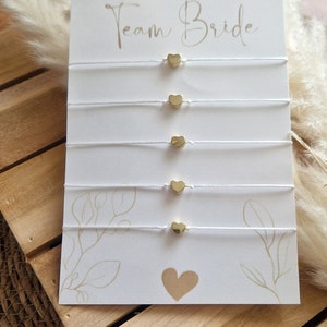 Könnte beinhalten: Eine weiße Karte mit dem Text "Team Bride" in schwarzer, kursiv geschriebener Schrift. Fünf weiße Schnüre mit goldenen Herzanhängern sind an der Karte befestigt.
