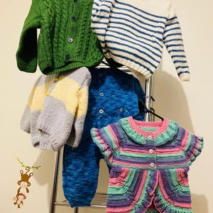Peut inclure: Plusieurs vêtements pour enfants tricotés à la main, dont des pulls et une combinaison, suspendus à un portant en métal. On distingue un cardigan vert en tricot torsadé, un pull rayé bleu et blanc et un cardigan multicolore à manches courtes.