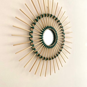 May include: A round mirror with a sunburst design made of natural wood sticks and green twine. The mirror is surrounded by a circle of wood sticks that are woven together with green twine.