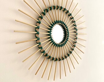 May include: A round mirror with a sunburst design made of natural wood sticks and green twine. The mirror is surrounded by a circle of wood sticks that are woven together with green twine.