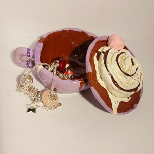 May include: A purple and brown ceramic trinket box shaped like a cup of hot chocolate with whipped cream and a cherry on top. The lid is open and a silver charm bracelet with beads is inside.