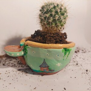 May include: A small green ceramic planter shaped like a teacup with a brown handle, painted with white dots and a small house design. A green cactus plant is growing in the planter.