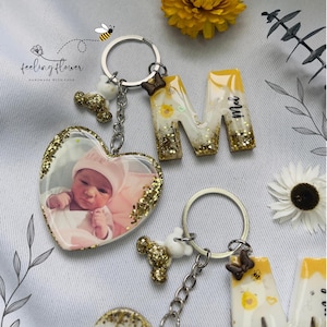 May include: A set of resin keychains with a heart-shaped charm featuring a baby photo, a letter M with glitter and the word "Mia", a bee charm, and a small bear charm. The keychains are attached to a silver chain and ring.