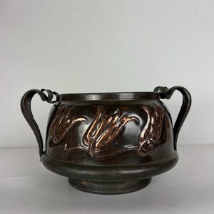 May include: A dark bronze decorative bowl with two curved handles. The bowl has a raised, stylized floral design in a lighter bronze colour around its circumference. The bowl sits on a circular base.