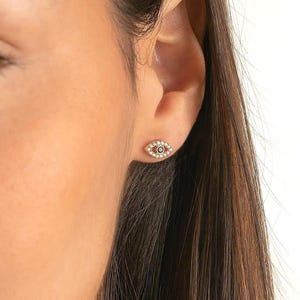 May include: Close-up of a person's face with an eye-shaped stud earring. The earring is gold-toned with a blue center and white accents. The earring is on the right ear. The person has brown hair and skin.