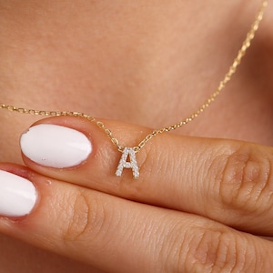 May include: A delicate gold necklace featuring a pendant in the shape of the letter "A", encrusted with small, sparkling gemstones. The necklace is displayed on a person's neck, with the chain and pendant clearly visible. The person's hand is gently holding the necklace.