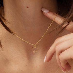 Op de afbeelding: Een delicate goudkleurige ketting met een Mickey Mouse-omtrek hanger. De hanger hangt aan een fijne ketting. De ketting wordt om de nek van een persoon gedragen, tegen een lichtroze top.
