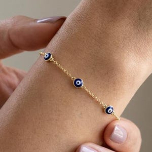 May include: A delicate gold chain bracelet featuring three evenly spaced circular charms. Each charm displays a dark blue, white, and black eye-shaped motif. The bracelet is secured with a thin, polished gold-tone metal link chain design.
