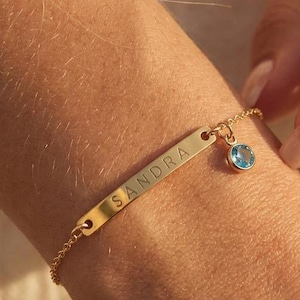 May include: A gold-colored bracelet with the name "SANDRA" engraved on a rectangular bar. A small, round, light blue gemstone charm hangs from the bar. The bracelet is on a wrist.