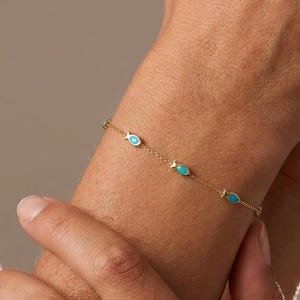 May include: A delicate gold chain bracelet featuring evenly spaced, small, oval-shaped turquoise-colored charms. The bracelet is constructed with a fine link chain and secures around the wrist, showcasing a minimalist design with vibrant blue accents.