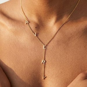 May include: A gold-colored necklace with the name "AVERY" spelled out in individual letters. The letters are connected by a delicate chain, with the "AV" portion of the name positioned to the left and the "ERY" portion hanging vertically.
