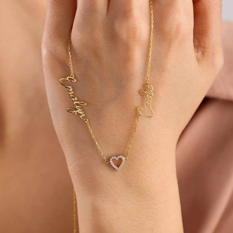 K&ouml;nnte beinhalten: Goldkette mit zwei Namen, "Cody" und "Emily", in Schreibschrift. Die Kette hat einen kleinen Herz-Anh&auml;nger mit einem diamantartigen Glanz.