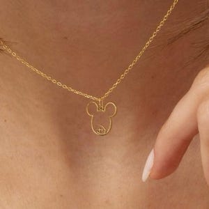 Op de afbeelding: Een delicate goudkleurige ketting met een hanger in de vorm van het hoofd van Mickey Mouse. De hanger is een eenvoudige omtrek met de iconische oren en gezichtsdetails. De ketting wordt om de nek van een persoon gedragen.