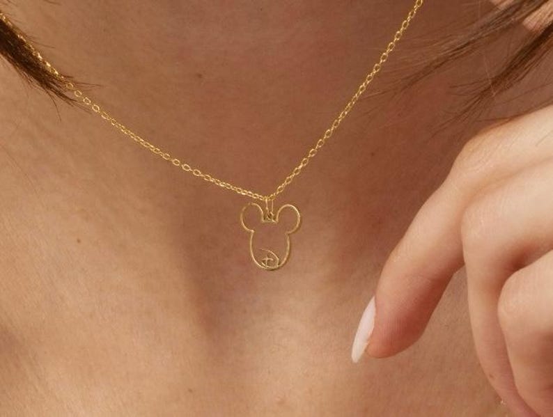 Op de afbeelding: Een delicate goudkleurige ketting met een hanger in de vorm van het hoofd van Mickey Mouse. De hanger is een eenvoudige omtrek met de iconische oren en gezichtsdetails. De ketting wordt om de nek van een persoon gedragen.