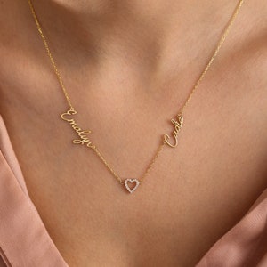 K&ouml;nnte beinhalten: Goldkette mit zwei Namen, "Cadence" und "Emilyn", in Schreibschrift. Ein kleiner Herz-Anh&auml;nger mit klaren Zirkonia-Steinen h&auml;ngt zwischen den Namen.