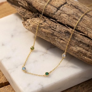May include: A delicate gold necklace featuring four colorful gemstones: a light blue, green, emerald green, and clear stone. The necklace is displayed on a white marble surface with a wooden background.