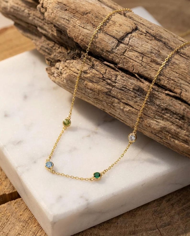 May include: A delicate gold necklace featuring four colorful gemstones: a light blue, green, emerald green, and clear stone. The necklace is displayed on a white marble surface with a wooden background.