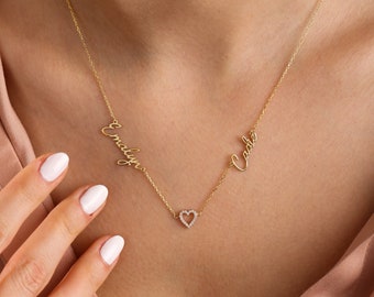 Könnte beinhalten: Goldkette mit zwei Namen, "Emilyn" und "Cadle", in Schreibschrift. Die Namen sind durch einen kleinen Herzanhänger mit einem diamantartigen Akzent verbunden.