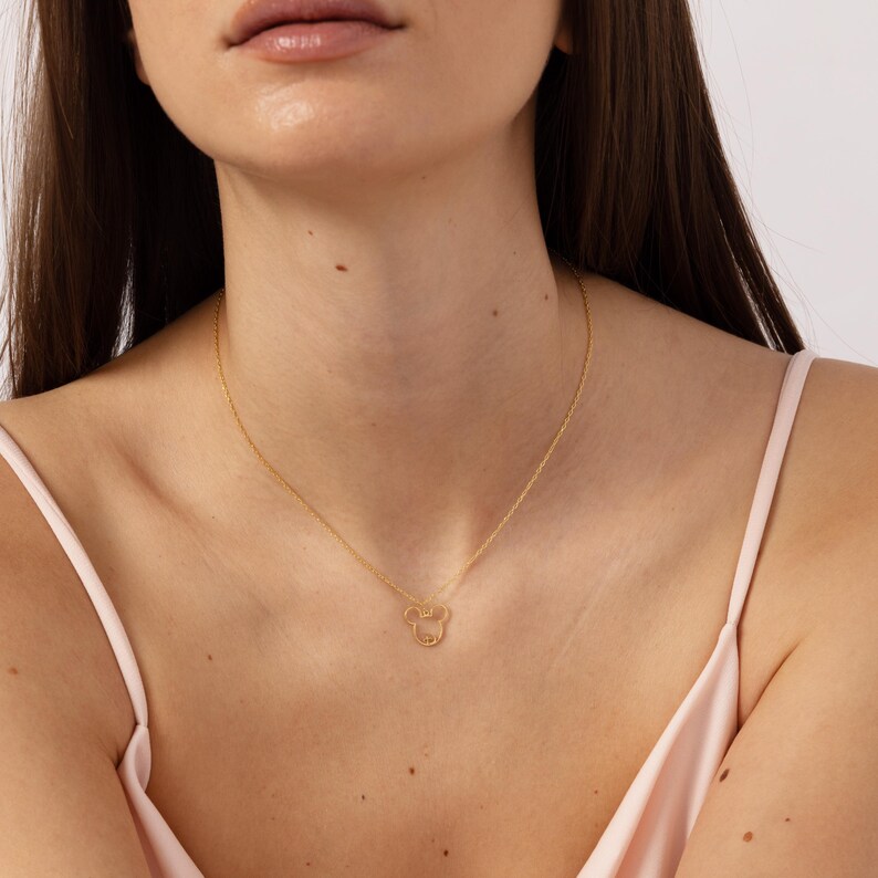 Op de afbeelding: Een goudkleurige ketting met een hanger in de vorm van een cartoon muizenkop. De hanger is een delicate omtrek met kleine details. De ketting wordt om de nek gedragen.