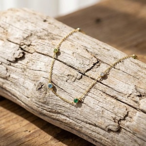 May include: A delicate gold necklace with several small, colorful gemstones. The necklace is draped across a piece of weathered wood, showcasing its fine chain and the vibrant hues of the stones, including green, blue, and clear gems.