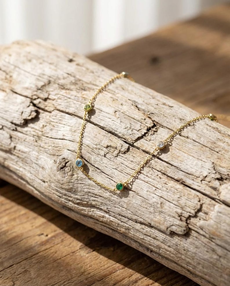 May include: A delicate gold necklace with several small, colorful gemstones. The necklace is draped across a piece of weathered wood, showcasing its fine chain and the vibrant hues of the stones, including green, blue, and clear gems.