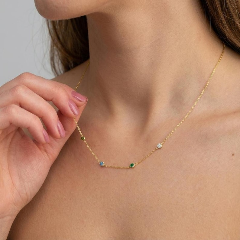 May include: A delicate gold necklace featuring a series of small, colorful gemstones. The necklace has a fine chain and includes a white, green, blue, and emerald-colored stone. The necklace is displayed against a skin tone background.