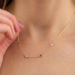 May include: A delicate gold necklace featuring five colorful gemstones: a light blue, green, white, and two olive-green stones. The necklace is displayed against a skin tone background, highlighting the jewelry's design and color.