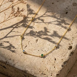 May include: A delicate gold necklace with several small, colorful gemstones. The necklace features a thin chain and is adorned with blue, green, and clear stones. The jewelry is displayed on a textured, light-colored surface.