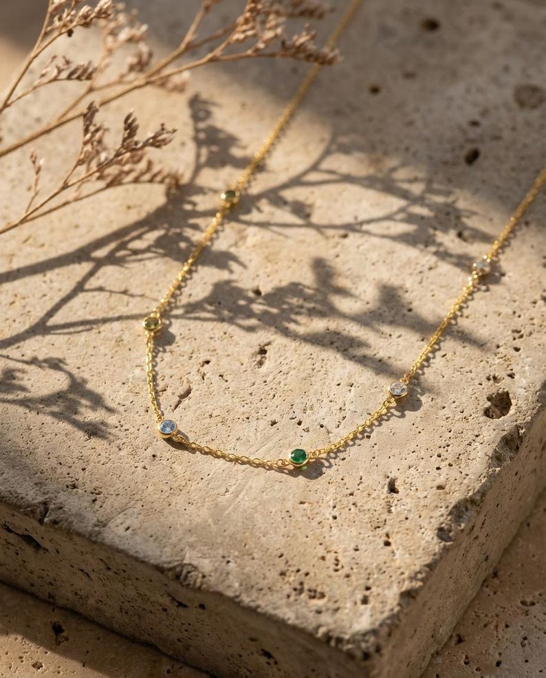 May include: A delicate gold necklace with several small, colorful gemstones. The necklace features a thin chain and is adorned with blue, green, and clear stones. The jewelry is displayed on a textured, light-colored surface.