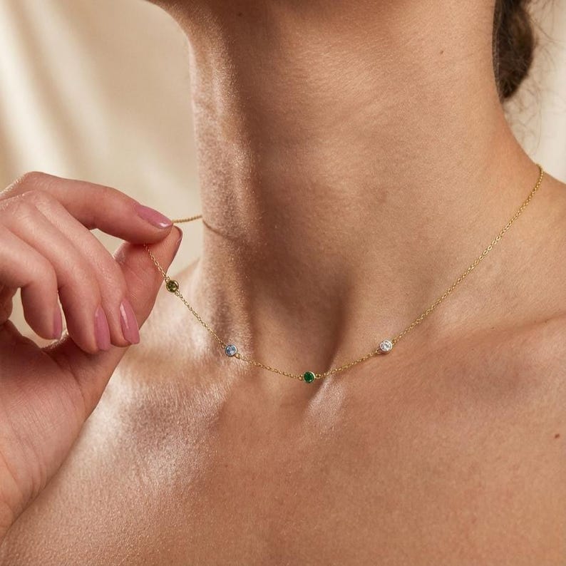 May include: A delicate gold necklace featuring small, colorful gemstones. The necklace has a fine chain and is adorned with a green, blue, white, and emerald-colored stone. The necklace is being held by a person.