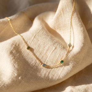 May include: A delicate gold necklace featuring five colorful gemstones: green, blue, emerald green, and clear. The necklace is displayed on a beige fabric, with the chain and gemstones catching the light.