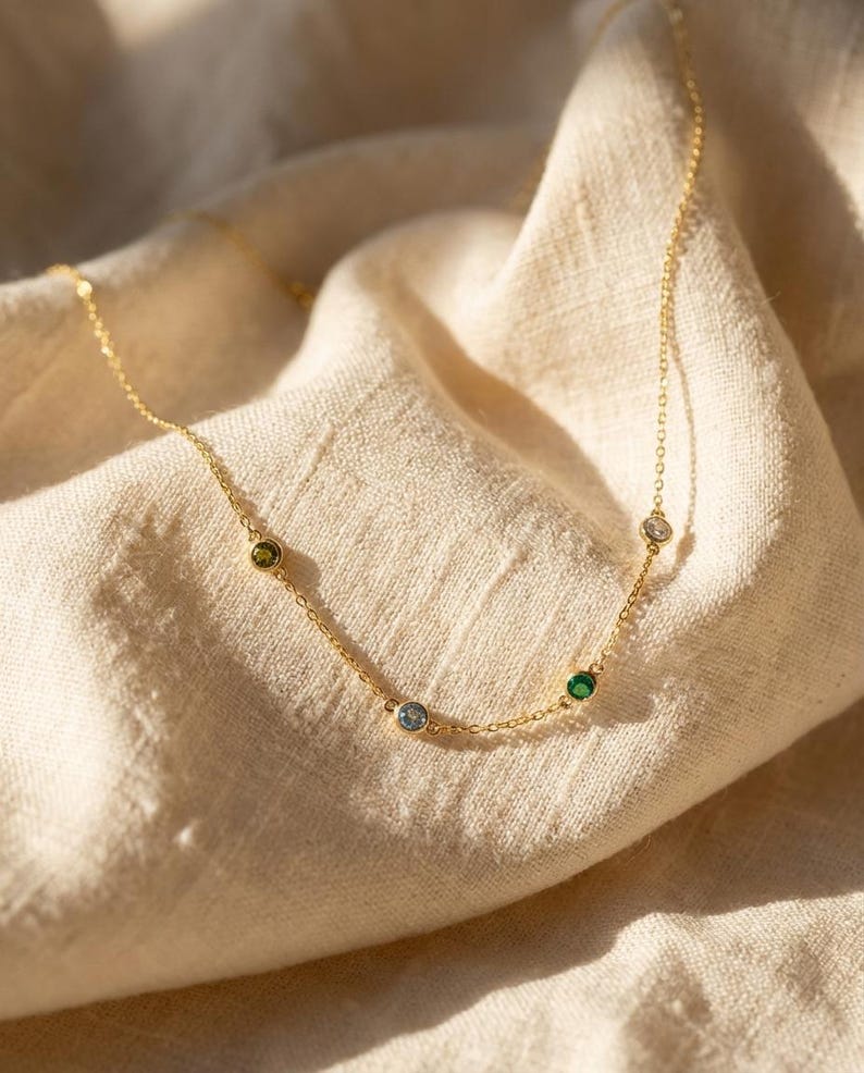 May include: A delicate gold necklace featuring five colorful gemstones: green, blue, emerald green, and clear. The necklace is displayed on a beige fabric, with the chain and gemstones catching the light.