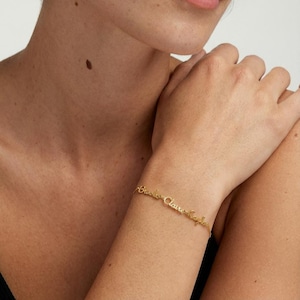 May include: A gold-colored bracelet with the names "Brooke Claire Taylor" elegantly inscribed. The delicate chain bracelet is worn on a wrist, showcasing the personalized jewelry. The names are in a cursive font.