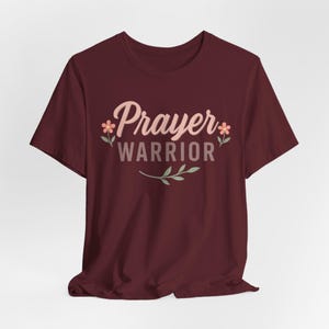 May include: A maroon t-shirt with the words "Prayer Warrior" in a light pink script font, accented with small flowers and a sprig of leaves. The shirt is made of a soft material and has short sleeves.