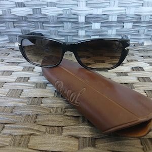 May include: Black sunglasses with dark gradient lenses and silver-tone accents on the temples, resting on a brown leather-like hard case embossed with the brand name Persol. The eyewear features a classic rectangular frame shape and dark arms.