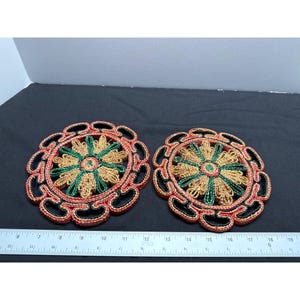 Puede incluir: Dos posavasos de paja trenzada con un diseño floral negro, rojo y verde. Los posavasos son redondos y miden aproximadamente 12,5 cm de diámetro.