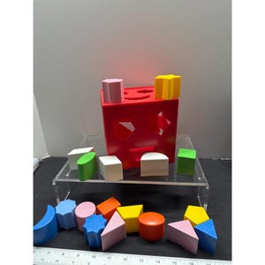 May include: A red shape sorter toy with various colorful wooden shapes. The shapes include cubes, cylinders, triangles, and other geometric forms. The sorter has openings on the top and sides to match the shapes. The toy is designed for early childhood development.