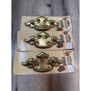 May include: Three brass-toned metal drawer pulls with a decorative backplate and a swinging bail handle. Each pull is mounted on a rectangular cardboard backing labeled with the brand name Belwith and includes two small metal mounting screws.