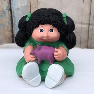 Statuetta Cabbage Patch Kids di una bambina che tiene in mano un salvadanaio - Dettagli vintage con vestito verde e fiocco