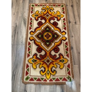 May include: A vintage rug with a colorful geometric pattern in shades of red, yellow, green, and brown. The rug has a cream-colored background and a brown border.