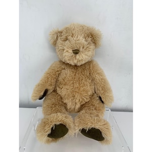 May include: A plush teddy bear with shaggy light brown fur, featuring a small dark brown nose and dark brown fabric patches on the soles of its feet and palms. The stuffed animal is seated in an upright position with its arms resting at its sides.