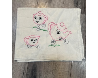 May include: A white tea towel with pink and green embroidered teapots and cups with faces. The teapots and cups have legs and are walking.