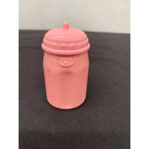 Puede incluir: Un pequeño recipiente de plástico rosa sólido con un cuerpo cilíndrico y una tapa de rosca a juego. La tapa presenta un borde festoneado texturizado, y la parte superior del recipiente muestra un pequeño detalle en relieve con forma de flor.