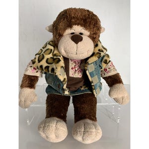 May include: A plush monkey toy with brown and beige fur, wearing a denim vest with a leopard print collar over a brown shirt featuring a pink lace-up detail and floral embroidery on the sleeves. The stuffed animal has a soft, rounded face.