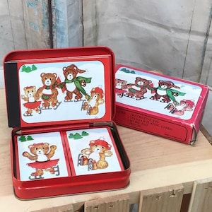 May include: A red metal tin containing two decks of playing cards, a score pad, and a pencil. The lid and card faces feature illustrations of brown teddy bears wearing winter clothing and ice skating in a snowy landscape with green pine trees.