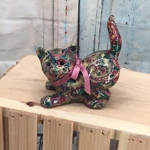 May include: A ceramic cat figurine featuring a colourful floral and paisley pattern in shades of red, green, yellow, and blue. The cat is in a crouching position with its tail held upright and is adorned with a light pink ribbon tied around its neck.