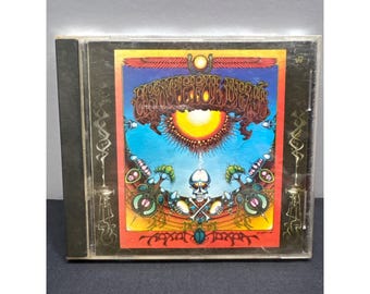May include: A CD case featuring the album art for the Grateful Dead's album, 'American Beauty'. The artwork is a vibrant mix of colors, including red, orange, blue, and green, with a skull and other psychedelic imagery. The CD case is in a clear plastic case.